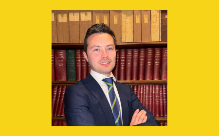 Michael Duggan stands in front of shelves of red and brown hardcover books, wearing a navy suit, white shirt, and striped tie, with a yellow background framing the image.